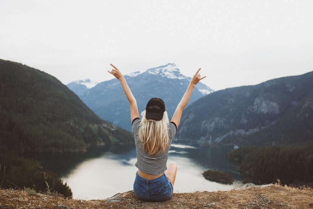 A blonde solo female traveller holds her hands up in excitement as she looks over a lake in a valley.